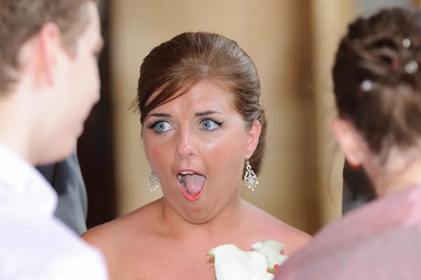 Woman with brown hair and earrings reacting with surprise, mouth open wide, holding a white flower.