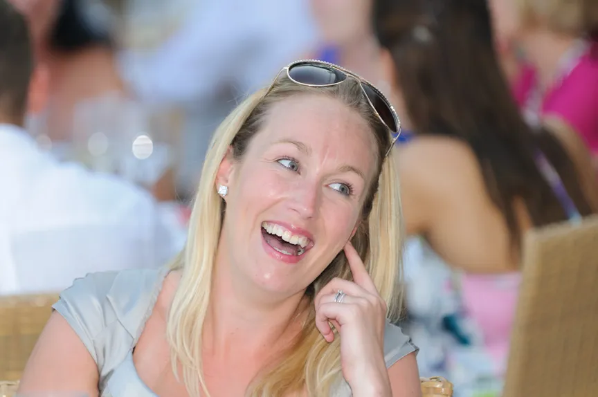 Blonde woman laughing with sunglasses on her head, seated in a crowded indoor setting.