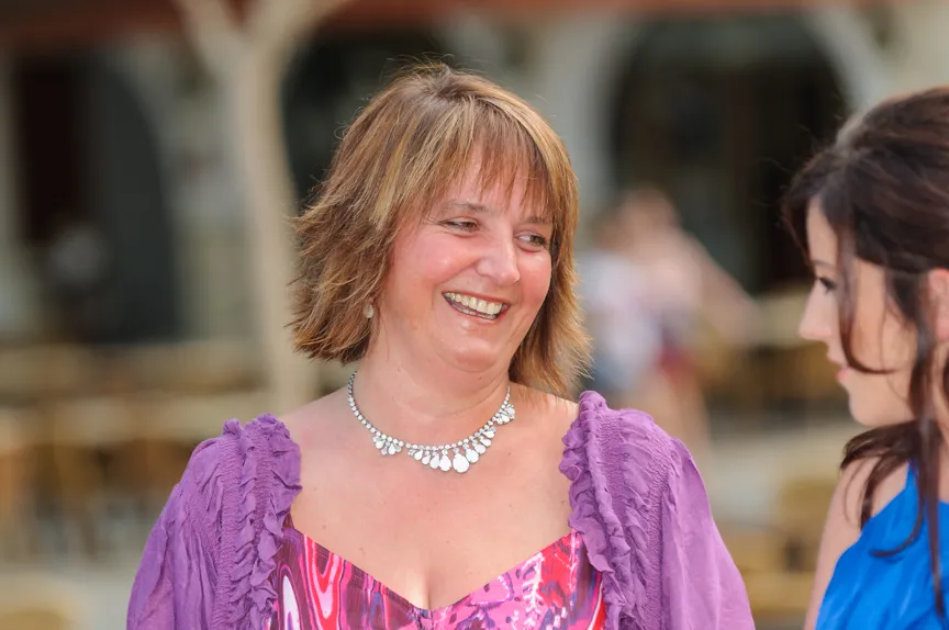 Smiling woman with short light brown hair wearing a purple dress and silver necklace, talking to another woman.