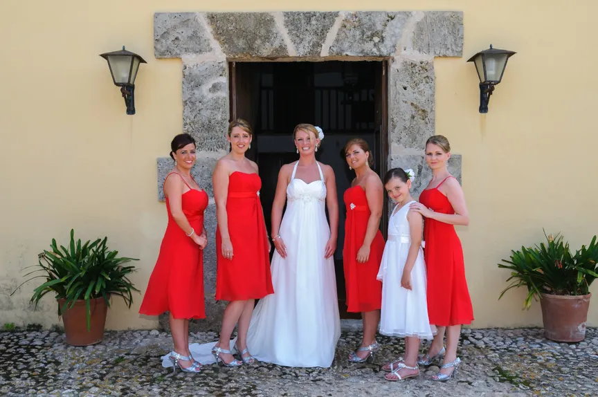 Bride in a white gown standing with five bridesmaids wearing red dresses and silver heels in front of a stone doorway.