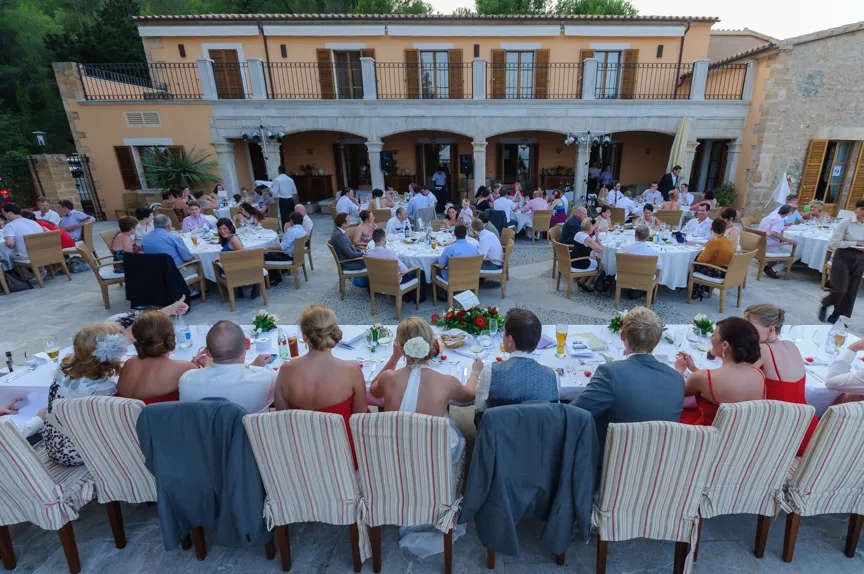 Outdoor wedding reception with guests seated at round tables and a long head table in front of a two-story building.