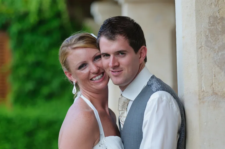 Smiling bride and groom embracing near a stone pillar with greenery in the background.