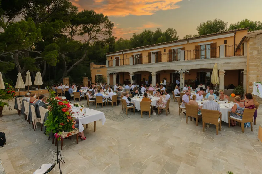 Guests seated at round tables with white tablecloths enjoying an outdoor dinner event at a villa during sunset.