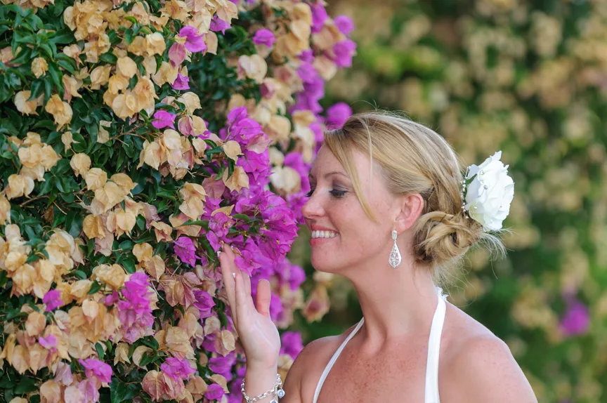 Smiling woman with blonde hair adorned with a white flower gently touching vibrant purple and beige bougainvillea flowers.