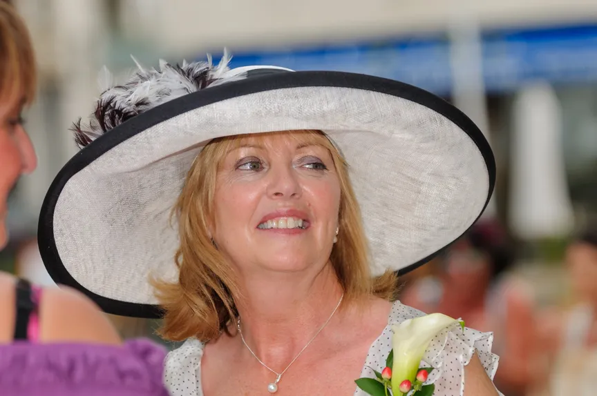 Smiling woman wearing a large white hat with black trim and a pearl necklace, holding a white calla lily corsage.