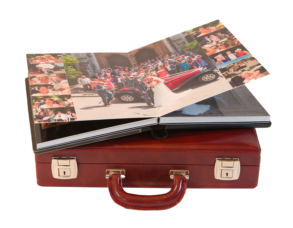 Open wedding photo album resting on a closed brown leather briefcase, showing a group photo with bride and groom by a vintage car.