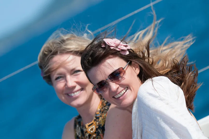 Two smiling women with windblown hair on a boat with a blue ocean background.