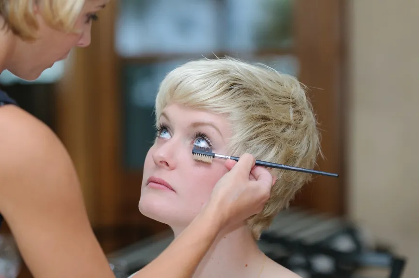 Makeup artist applying mascara to a woman's lower eyelashes with a brush.