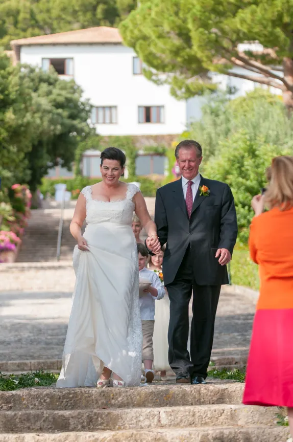 Bride in white dress walks down stone steps holding hands with a man in a dark suit, outdoors with greenery and buildings in the background.