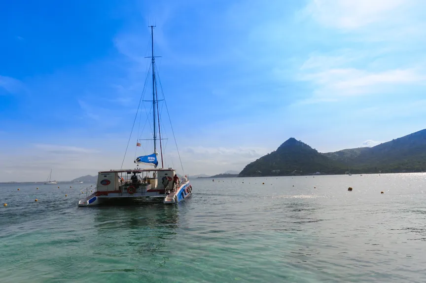 Catamaran with passengers sailing on calm blue water near a mountainous coastline under a partly cloudy sky.