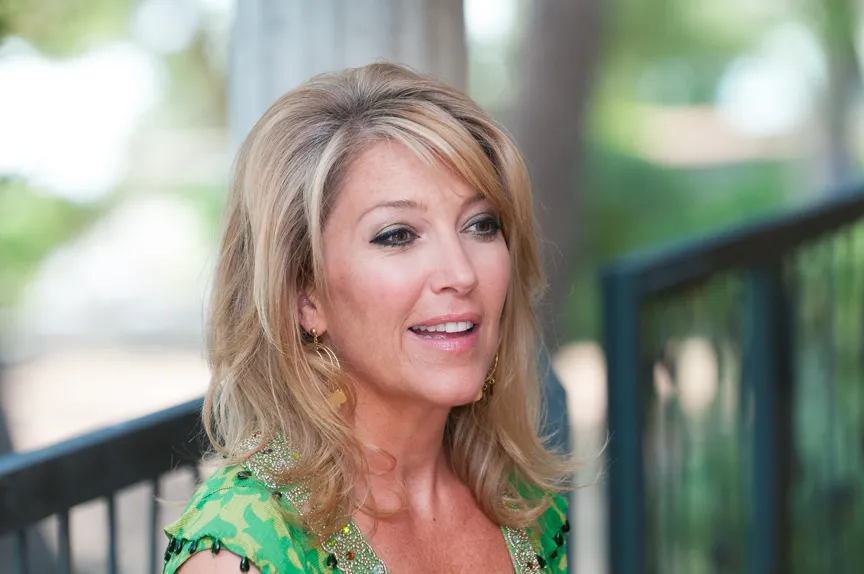 Blonde woman in a green dress with decorative trim, smiling softly while looking right, outdoors with blurred background.