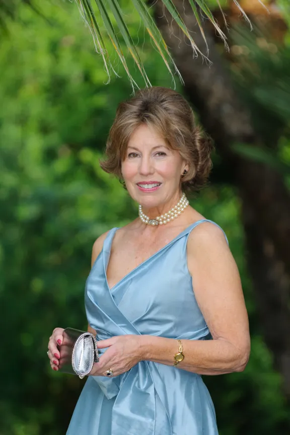 Smiling woman in light blue satin dress holding a silver clutch, standing outdoors with green foliage background.