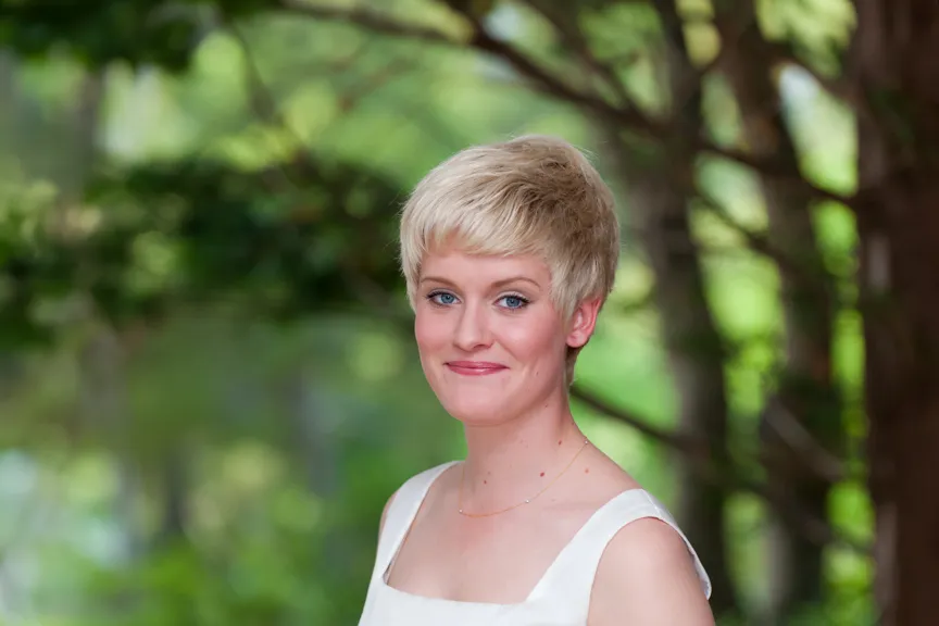 Smiling woman with short blonde hair wearing a white sleeveless top in a green outdoor setting.