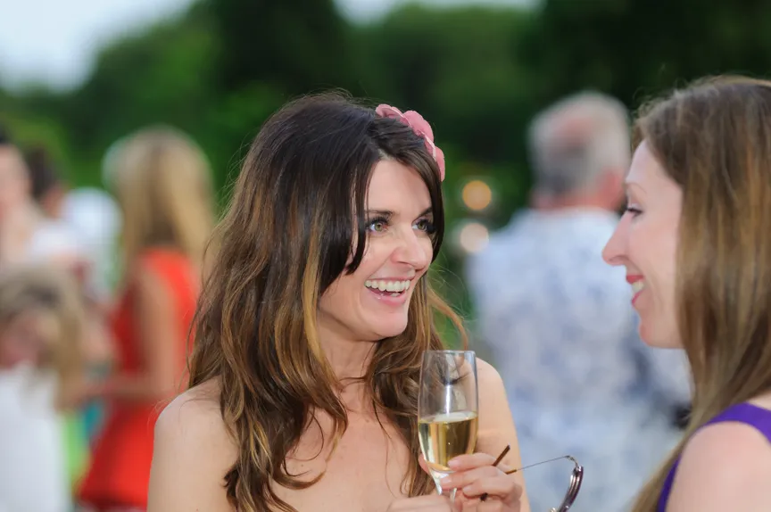 Two women smiling and talking, one holding a glass of champagne and a pair of sunglasses at an outdoor gathering.