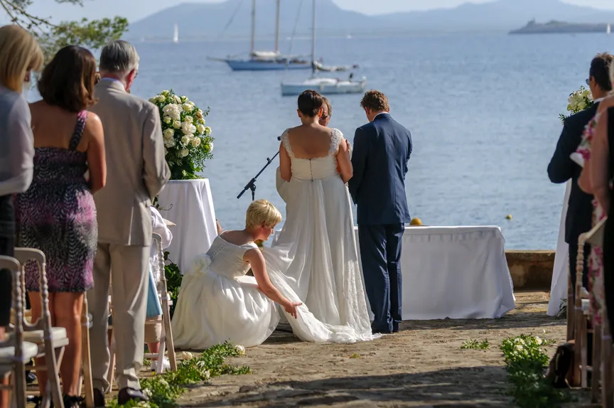 Outdoor wedding ceremony by the sea with bride, groom, and bridesmaid adjusting bride's dress train.