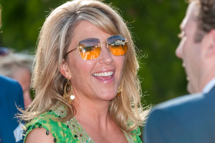 Smiling woman with blonde hair wearing orange-tinted sunglasses and a green dress talking to a man.