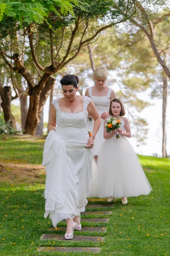 Bride in white dress walking down grassy steps followed by two girls in white dresses, one holding a flower bouquet.