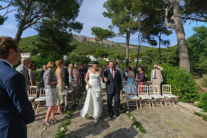 Bride in white wedding dress walking arm-in-arm with a man down an outdoor aisle lined with guests and chairs under tall trees.