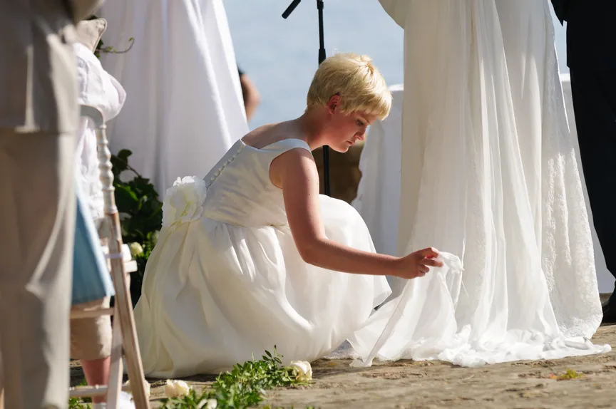 Young girl in a white dress crouching to adjust the bride's flowing wedding gown train during an outdoor ceremony.