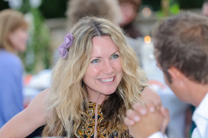 Smiling blonde woman with a purple flower in her hair talking to a man at a social gathering.