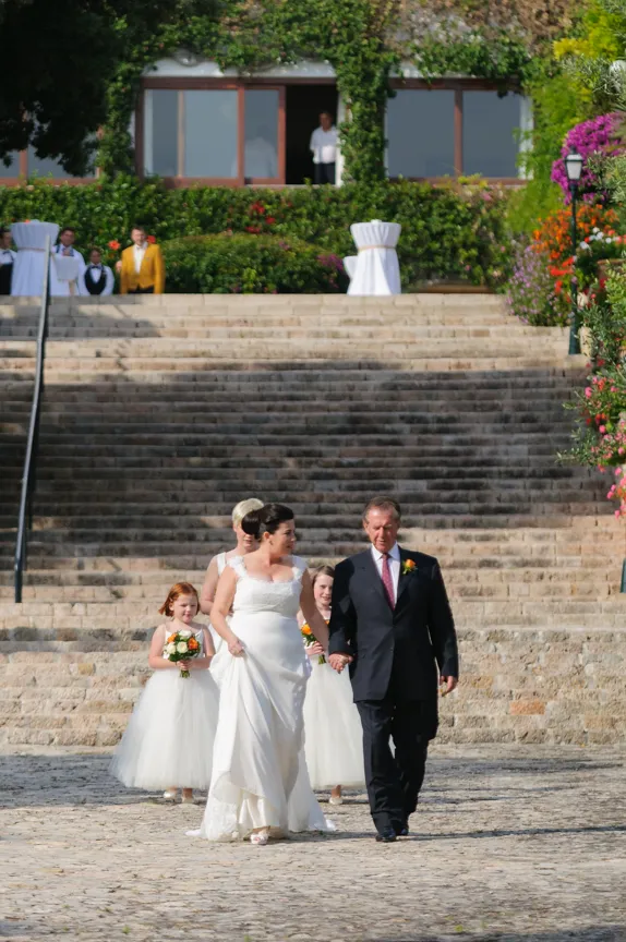 Bride in white dress walking down stone steps holding hands with a man in a suit, followed by two flower girls in white dresses holding bouquets.