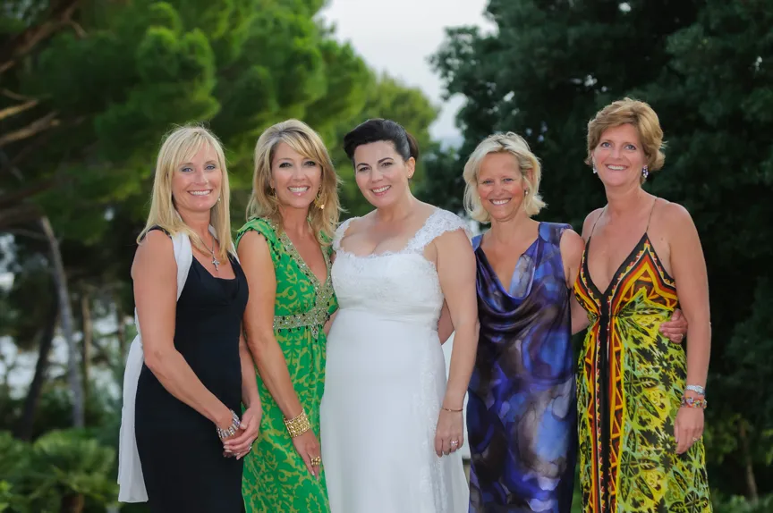 Five women standing outdoors smiling, with the center woman in a white wedding dress and the others in colorful dresses.