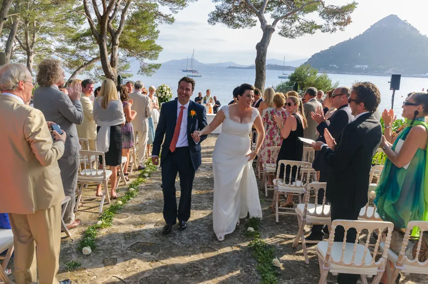 Bride and groom holding hands walking down an outdoor stone aisle by the sea as guests stand and applaud.