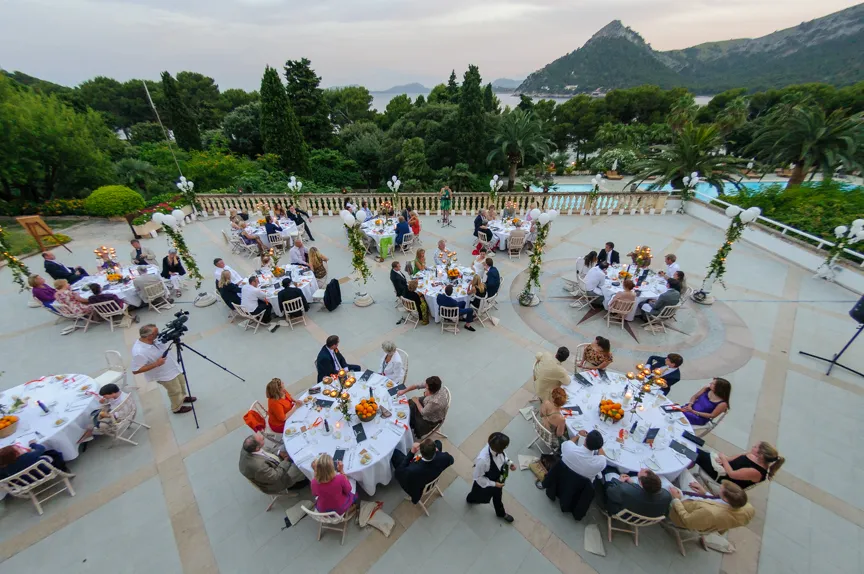 Outdoor formal dinner event with round tables, guests seated, elegant decorations, and a scenic mountain and sea background.
