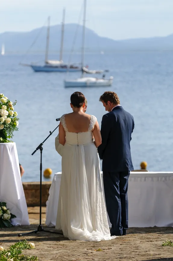 Bride in white dress and groom in navy suit standing together near a microphone by the water with sailboats in the background during an outdoor wedding ceremony.
