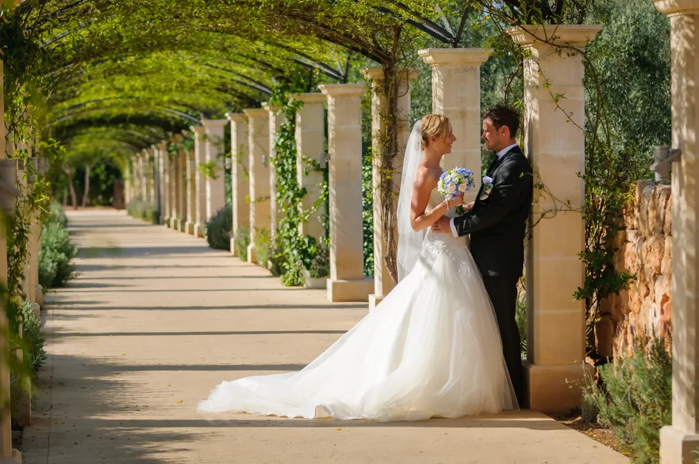 Bride and groom smiling and holding hands under a green vine-covered archway on a sunny day.