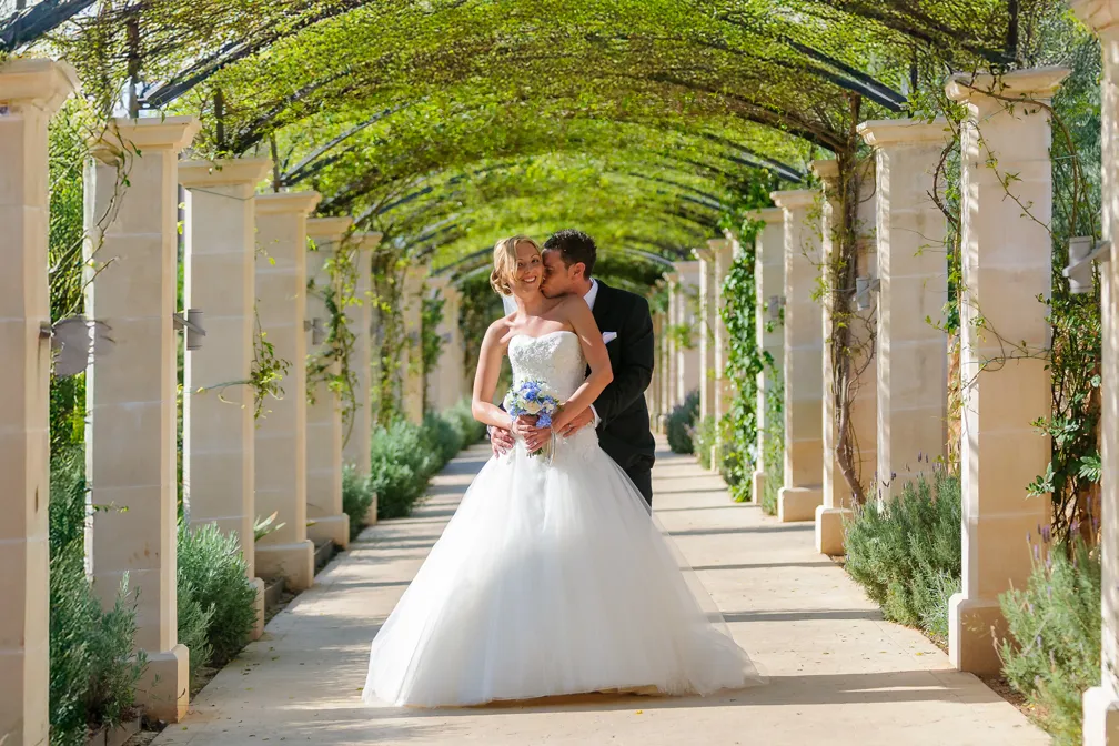 Bride in a white strapless gown holding a bouquet, embraced and kissed on the cheek by groom in a black suit, under an archway with green foliage vines.