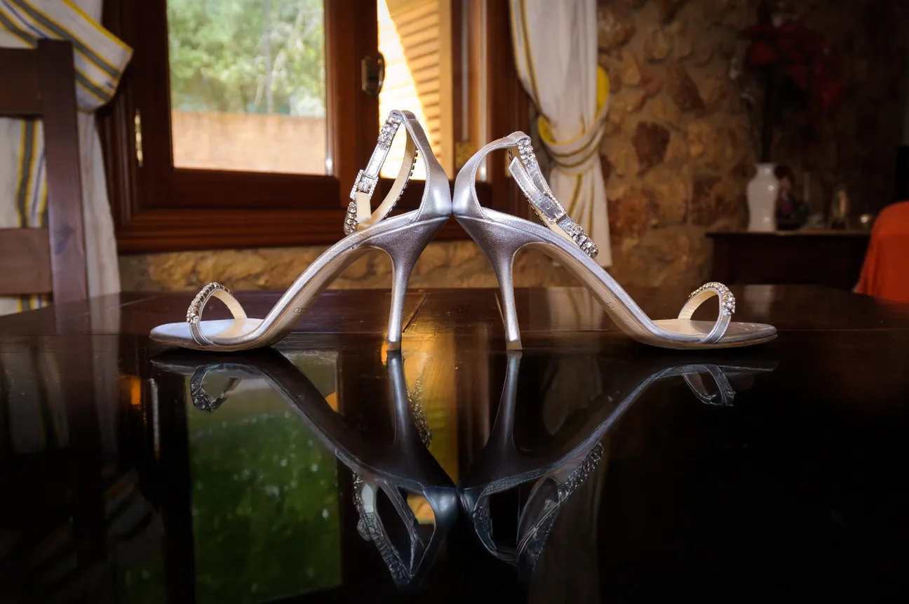 Pair of silver high-heeled sandals with rhinestone straps placed heel-to-heel on a reflective dark surface.