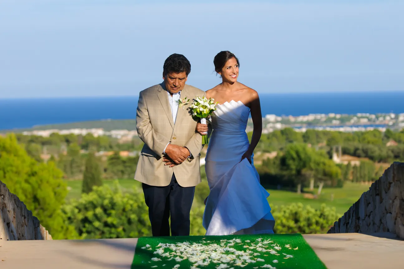 Bride in a white strapless gown holding a bouquet and walking arm-in-arm with a man in a beige suit down an aisle covered with white flower petals, with a scenic ocean and greenery background.