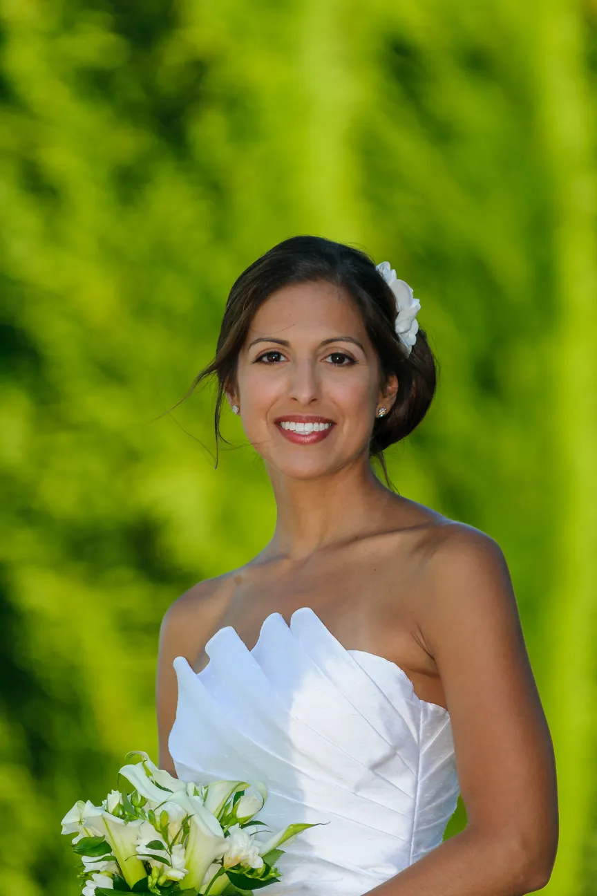 Smiling bride in a white strapless wedding dress holding a bouquet of white calla lilies with a blurred green background.