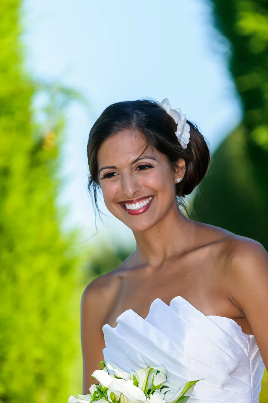 Smiling bride with a white flower in her hair holding a bouquet of white flowers outdoors.