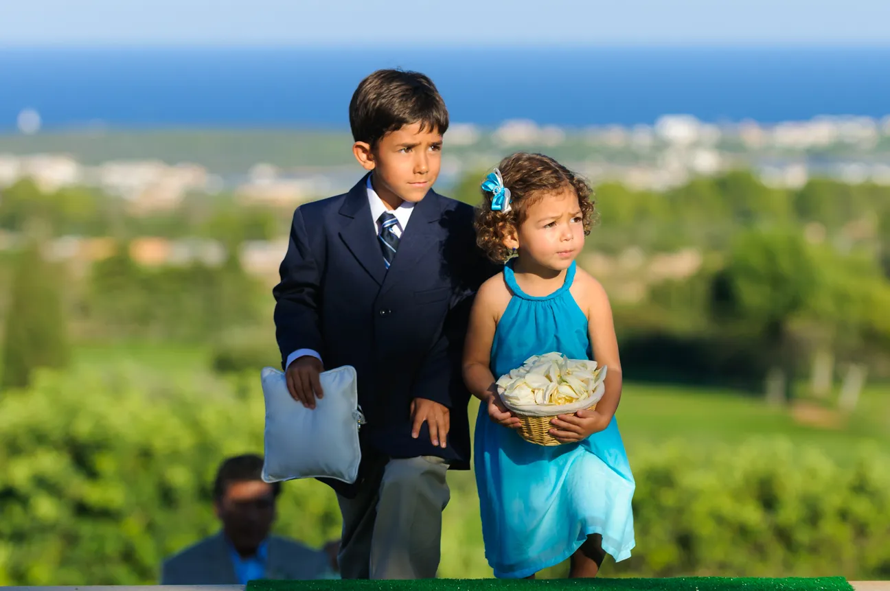 Young boy in a suit holding a white pillow and girl in a blue dress holding a basket of white rose petals outdoors.