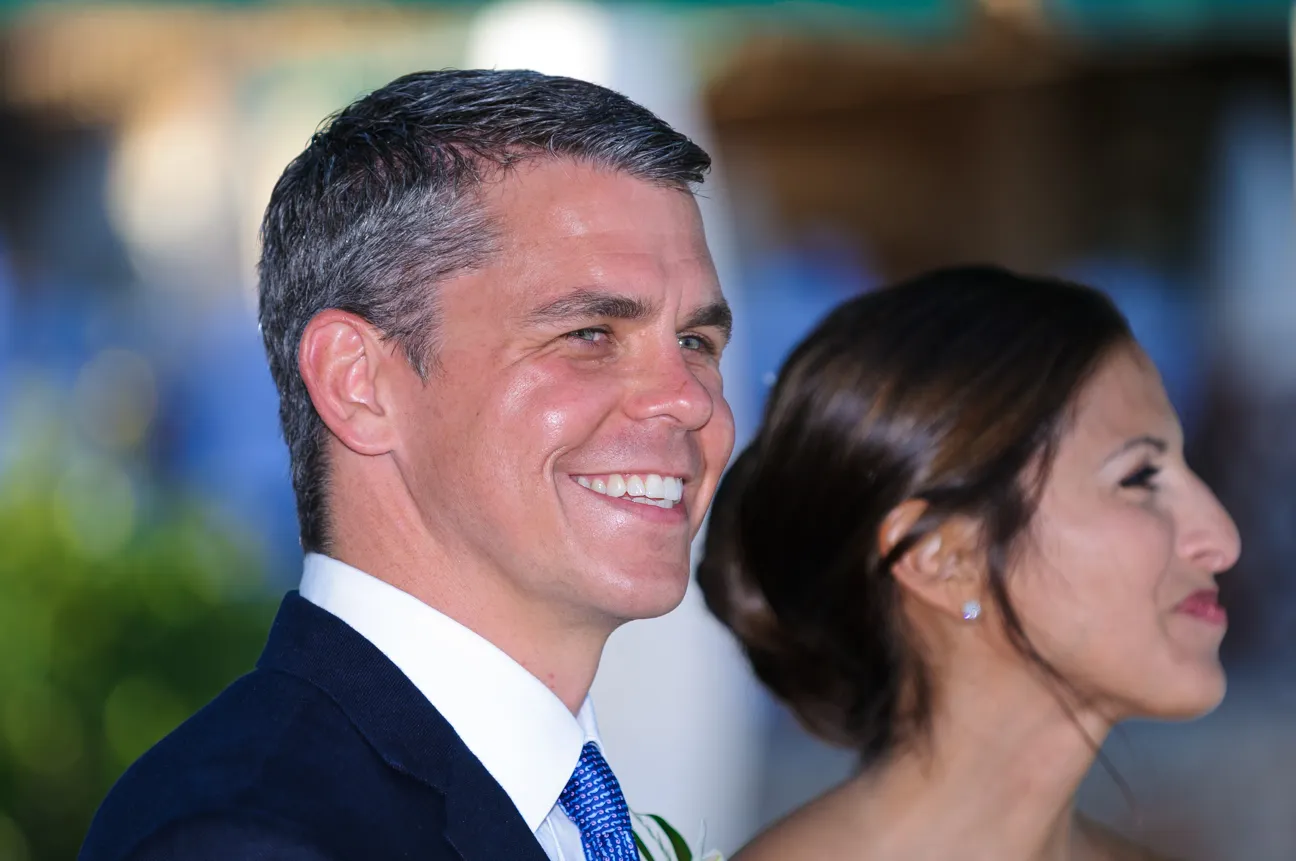 Close-up of a smiling man in a dark suit and blue tie with a woman in profile beside him.