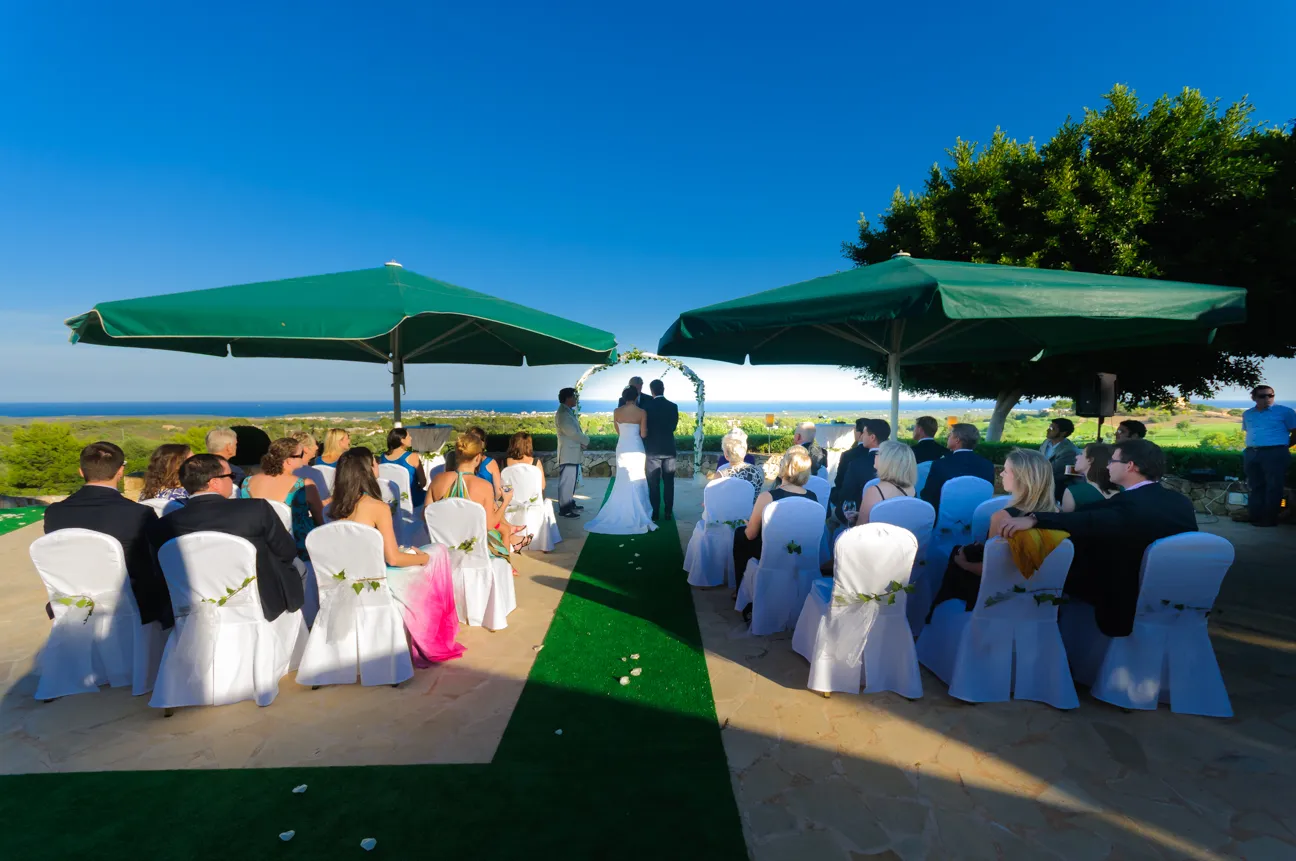 Outdoor wedding ceremony with bride and groom standing under a floral arch and guests seated on white-covered chairs beneath green umbrellas.