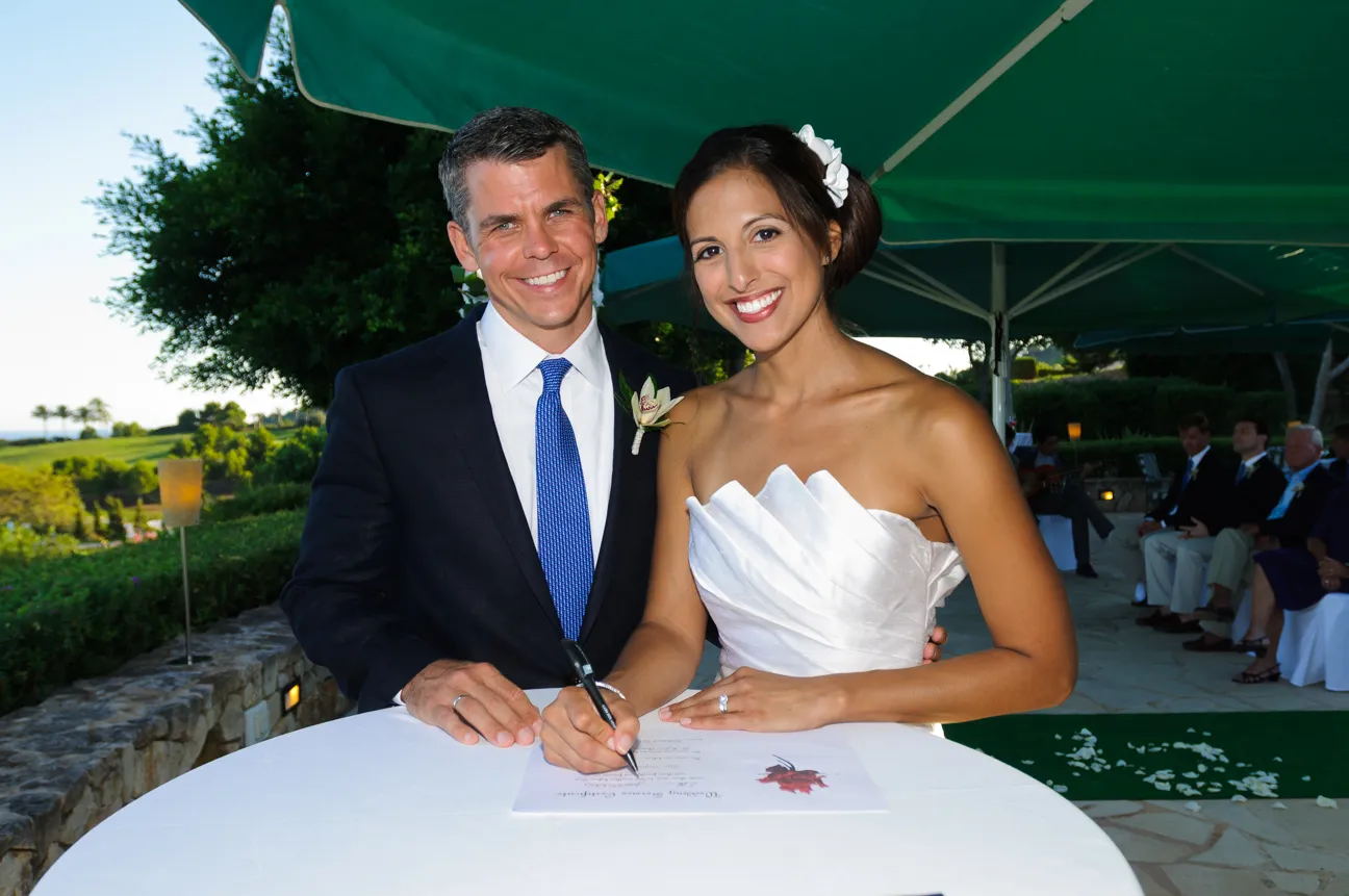 Bride in white wedding dress and groom in dark suit smiling as bride signs a document on a table outdoors.