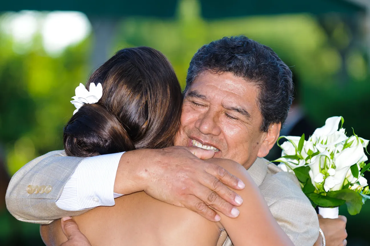 Man warmly hugging a woman with a white flower in her hair and holding a bouquet of white flowers.