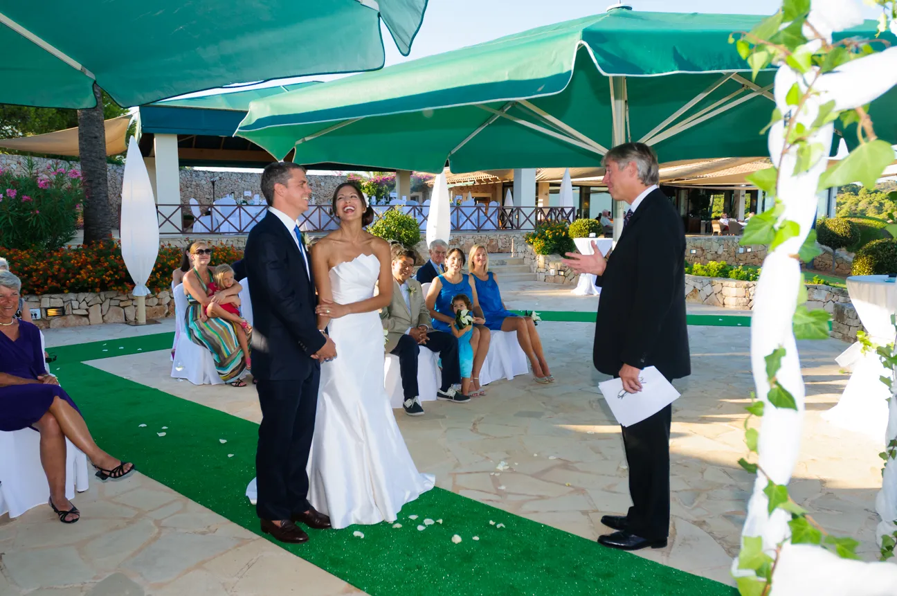 Bride and groom standing under green umbrellas during an outdoor wedding ceremony with guests seated nearby.