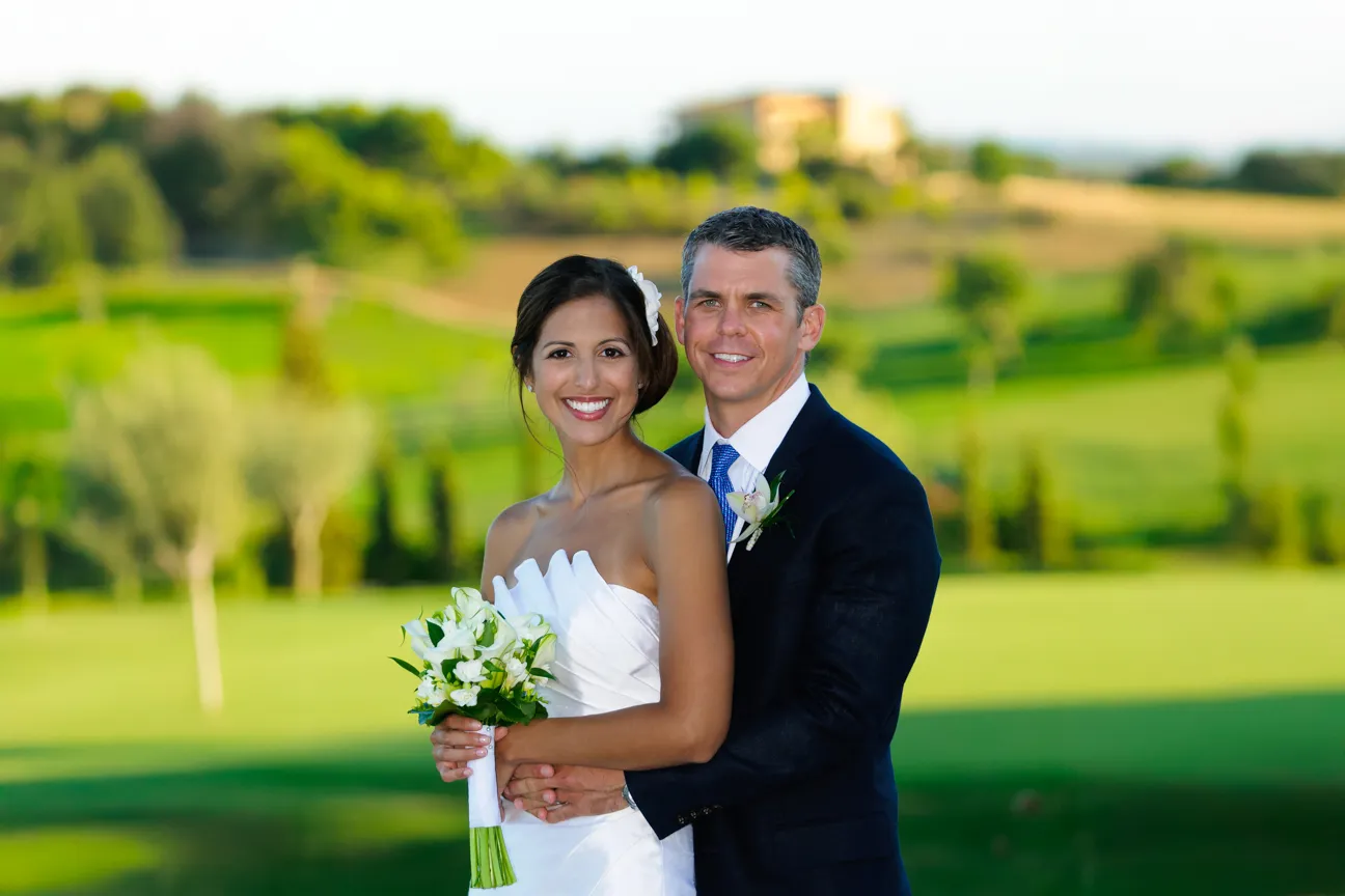Smiling bride in white dress holding bouquet and groom in dark suit embracing outdoors on a sunny day.