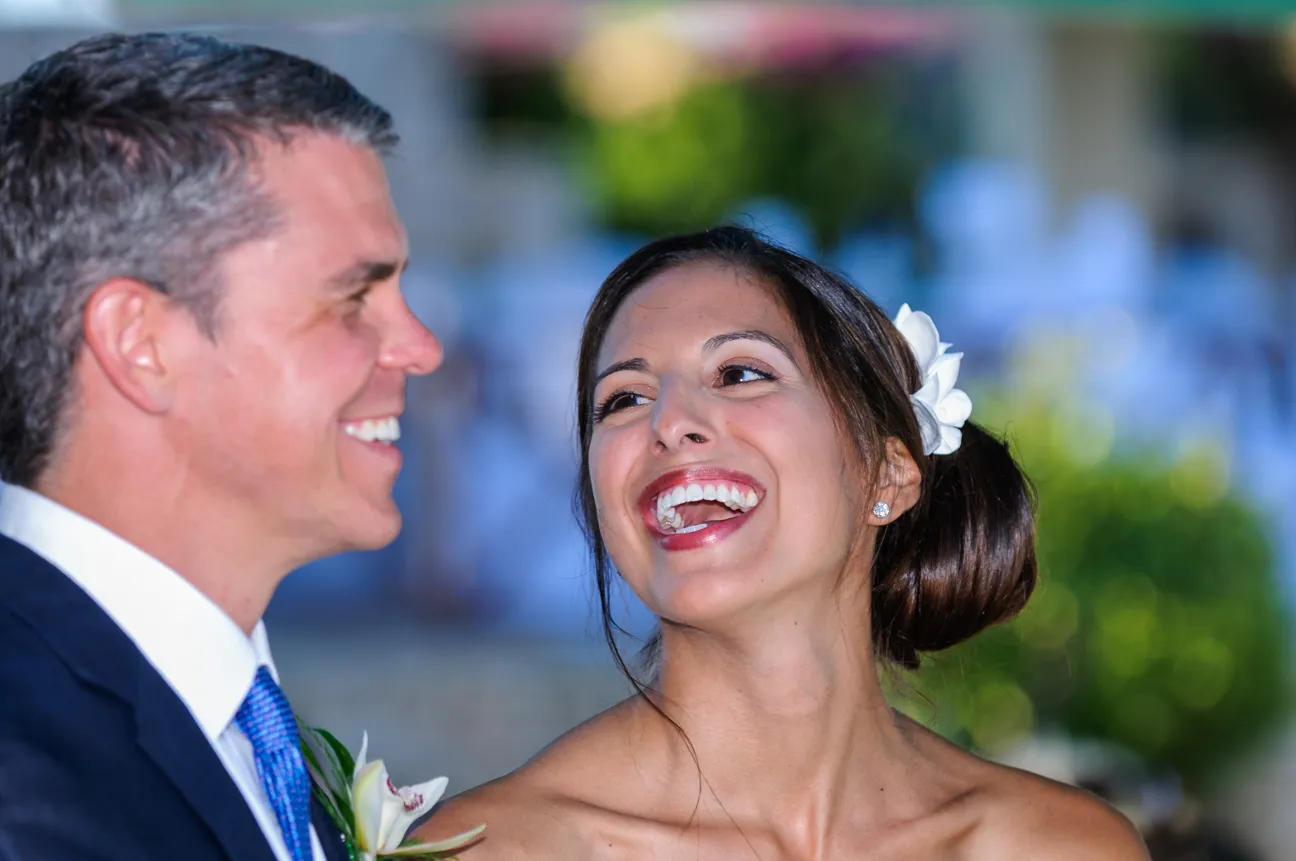 Smiling bride with a white flower in her hair looking at the groom, who is wearing a suit and tie, at an outdoor wedding.