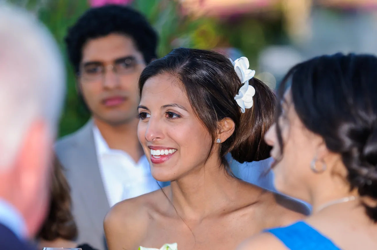 Smiling bride with white flowers in her hair talking to guests at an outdoor event.