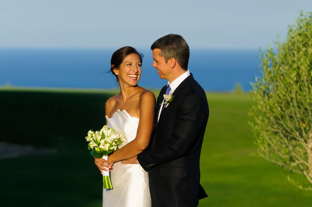 Smiling bride in white strapless dress holding a bouquet and groom in black suit embracing her outdoors with ocean and green grass in the background.