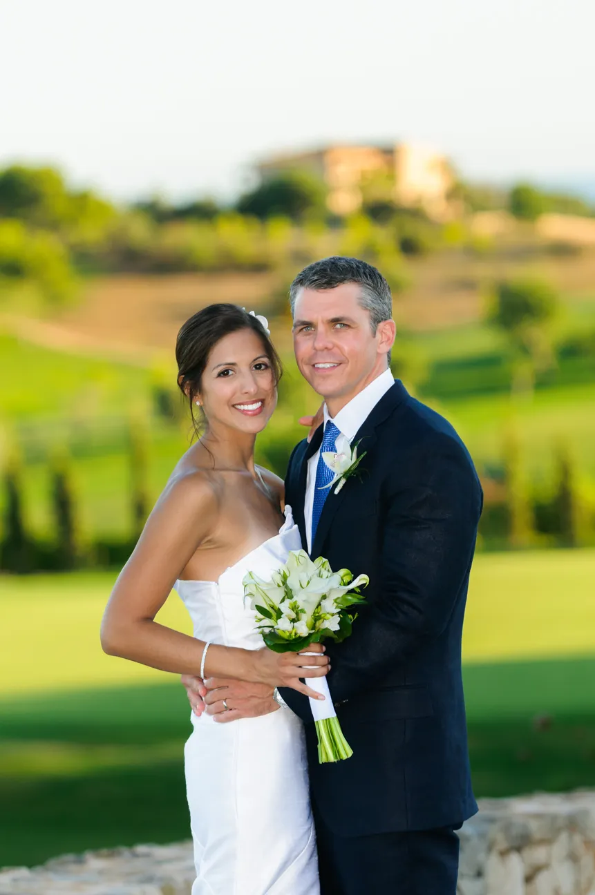 Bride in white dress and groom in dark suit embracing outdoors with green landscape background.