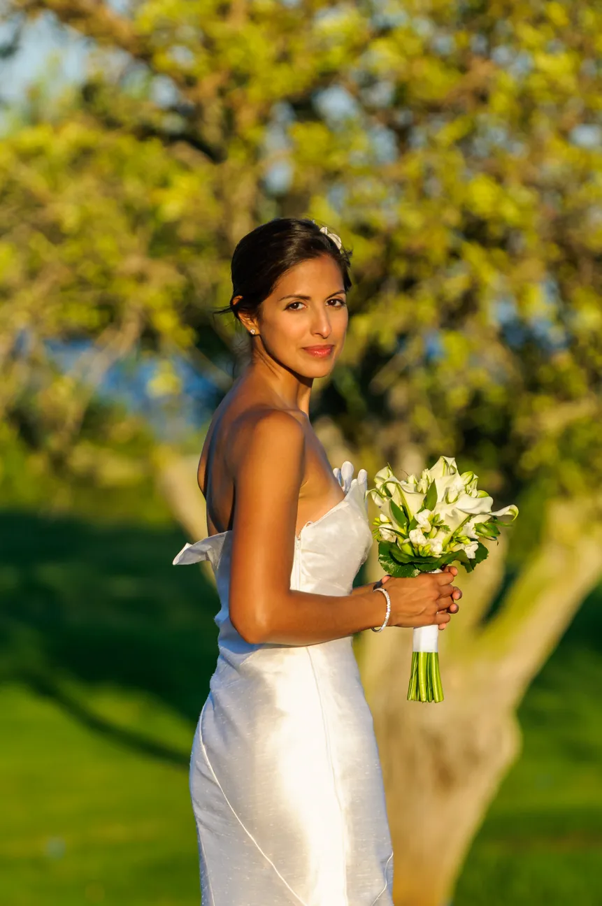 Bride in a white strapless wedding dress holding a bouquet of white flowers outdoors in sunlight.