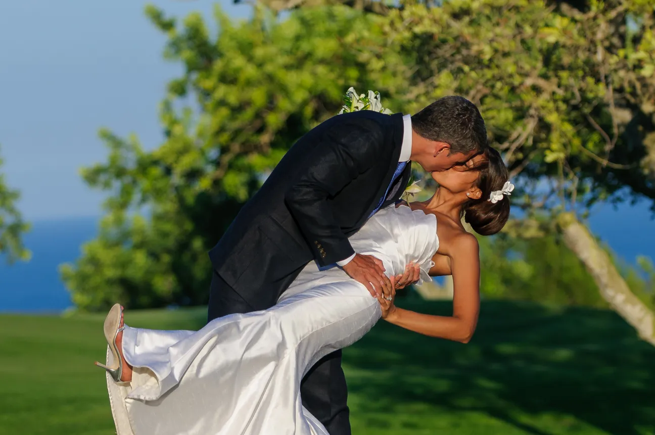 Groom in a dark suit dipping and kissing bride in a white wedding dress outdoors with green trees and blue sky background.