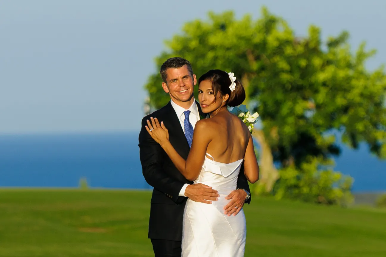 Bride and groom embracing outdoors with green grass, tree, and ocean in the background.
