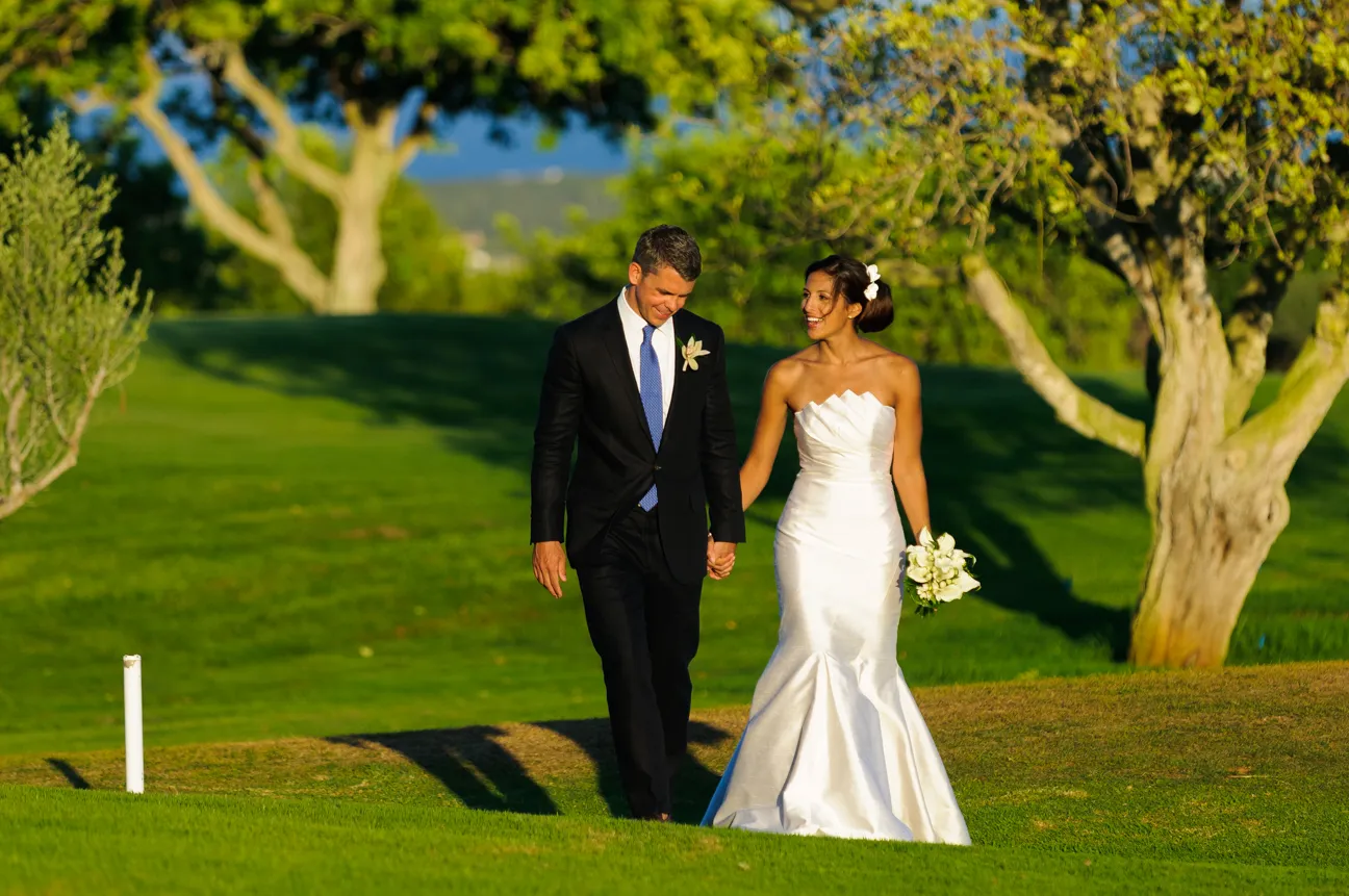 Bride in white strapless gown holding bouquet and groom in black suit walking hand in hand on green lawn with trees.
